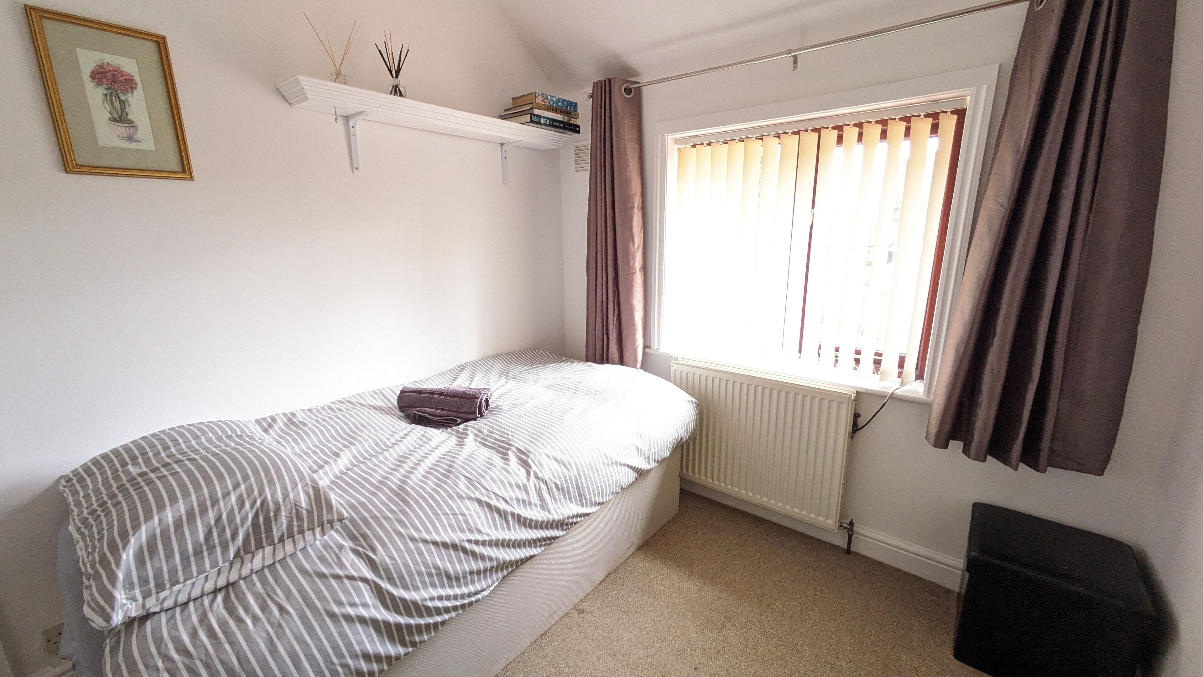 A small, single-bed room with a radiator, a window with curtains, a shelf with books and reed diffusers, and a framed picture on the wall.