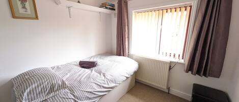 A small, single-bed room with a radiator, a window with curtains, a shelf with books and reed diffusers, and a framed picture on the wall.