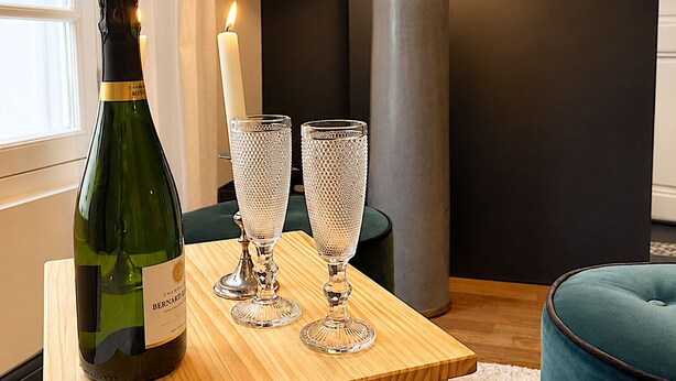 Et træbord med en vinflaske, to glas, et stearinlys og en gulvlampe i et rum med en mørk væg og et vindue.