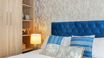 Una cama bien hecha con un cabecero azul, dos almohadas estampadas, una almohada decorativa y una lámpara de noche. Se ve un armario de madera a la izquierda.