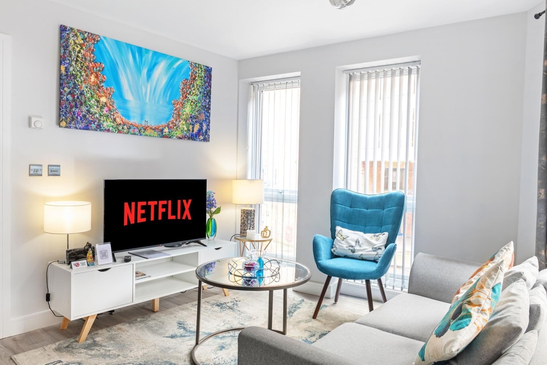 A modern living room with a flat-screen TV, a blue armchair, a grey sofa, and a colorful abstract painting on the wall.