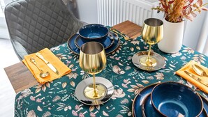 A dining table set with blue and gold dinnerware, a vase with dried flowers, and a chair with a quilted backrest.