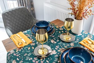 Una mesa de comedor dispuesta con vajilla azul y dorada, un florero con flores secas y una silla con respaldo acolchado.