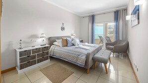 A modern hotel room with a bed, bedside tables, a small desk, a chair, and a balcony with curtains.
