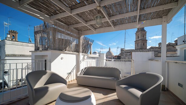 An outdoor seating area with modern furniture, a covered roof, and a view of a historic building in the background. - Le Contrade Chiasso Cafaro (Monopoli)