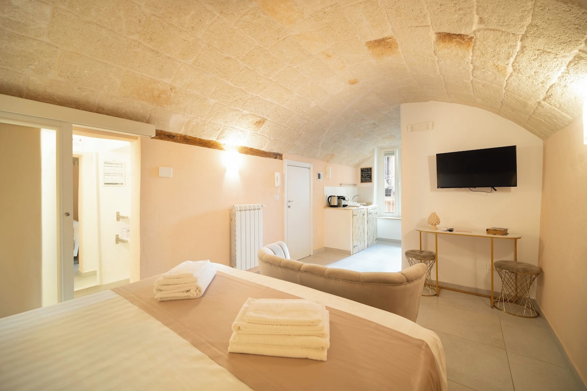 A room with a bed, a chair, a table, a TV, and a kitchen area in the background.
