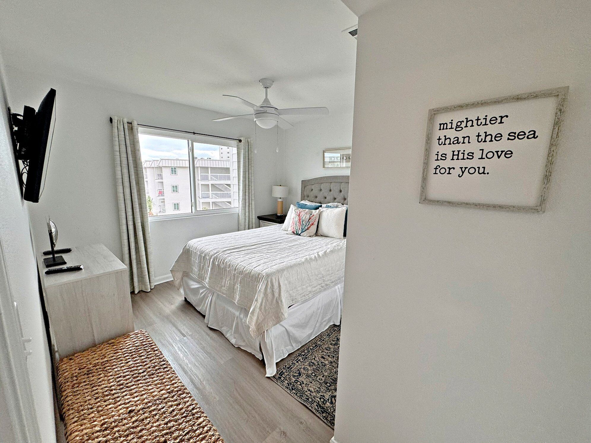 A bedroom with a bed, a ceiling fan, a bedside table, a nightstand, a window with curtains, and a motivational quote on the wall.