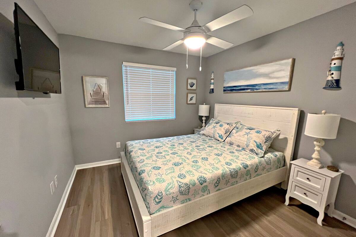 A bedroom with a bed, a ceiling fan, a nightstand, a lamp, a painting, and a window with blinds.
