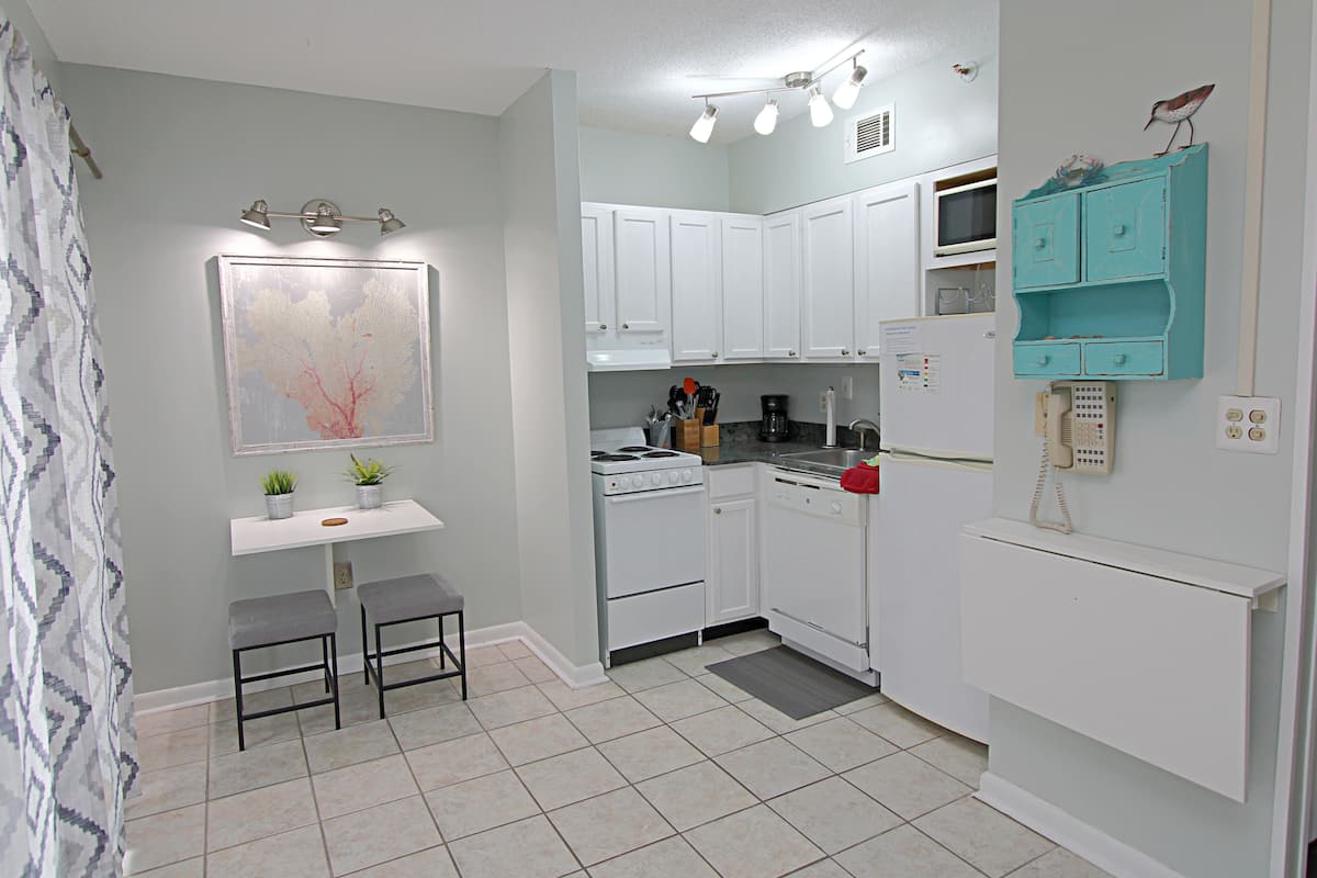 A compact kitchen with white cabinets, a stove, a microwave, and a teal-colored storage unit. There are two stools and a small table with plants.