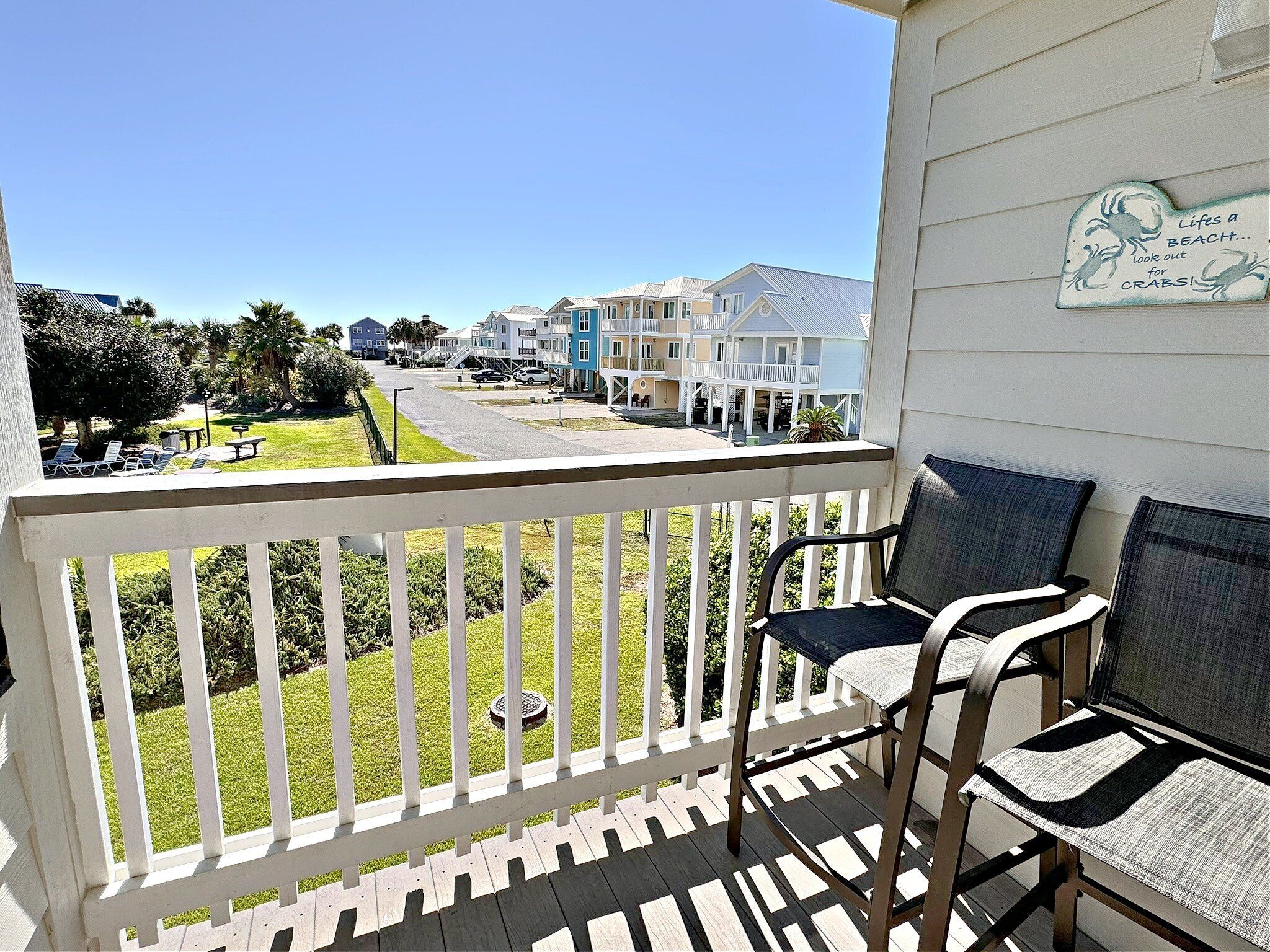 A balcony with white railings, two chairs, and a view of a beachfront community with houses and a clear sky.