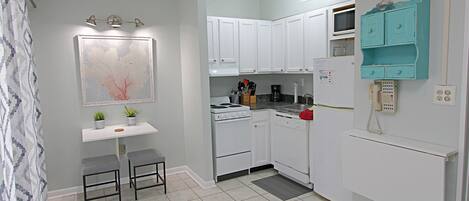 A compact kitchen with white cabinets, a stove, a microwave, and a teal-colored storage unit. There are two stools and a small table with plants.