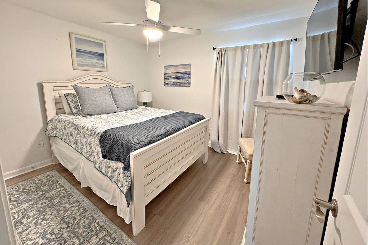 Ein Schlafzimmer mit einem Bett, einem Deckenventilator, einem Nachttisch, einer Lampe, einem kleinen Tisch mit einem dekorativen Gegenstand und einer Tür.