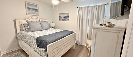 A bedroom with a bed, a ceiling fan, a nightstand, a lamp, a small table with a decorative item, and a door.