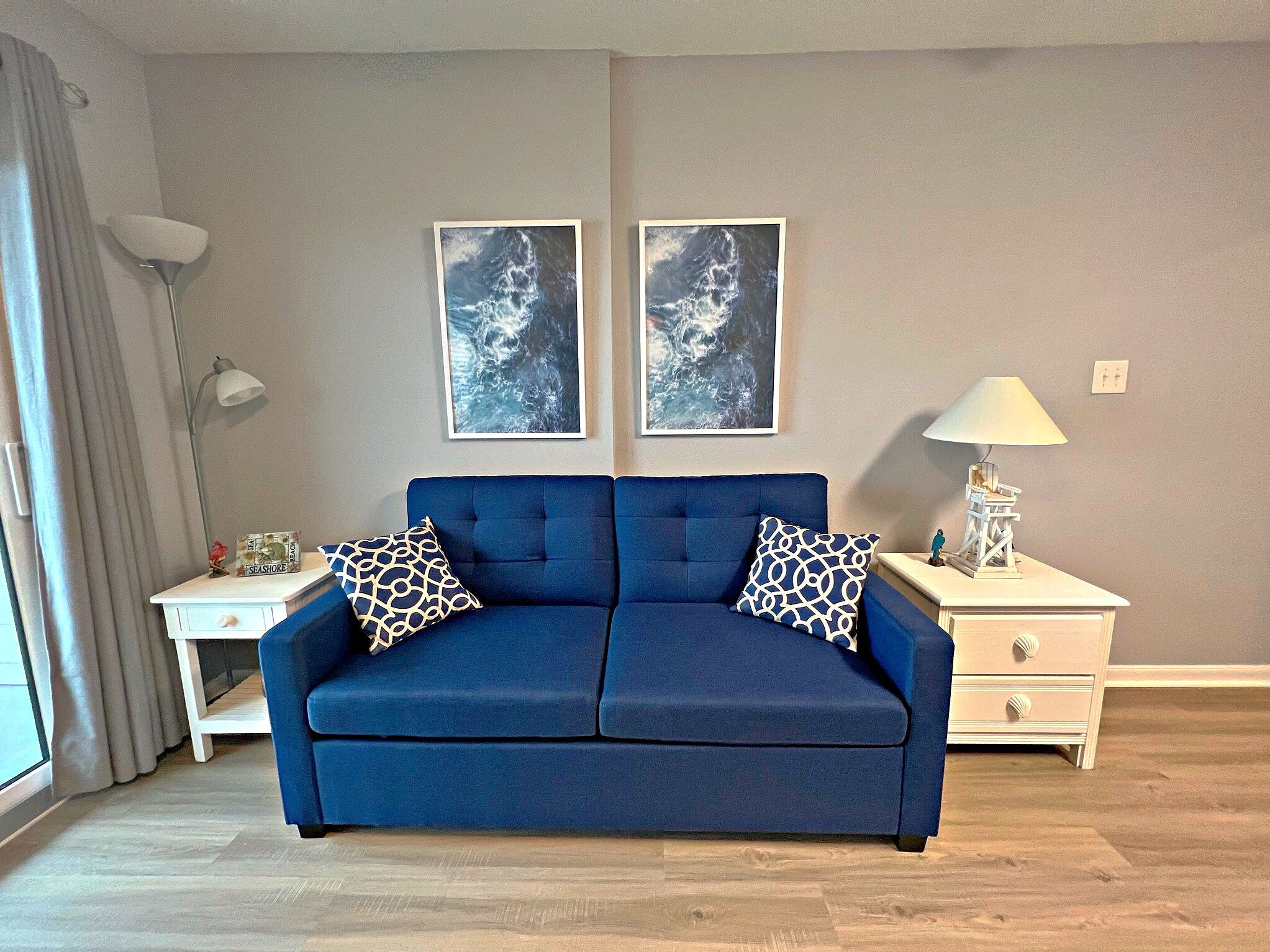 A blue sofa with patterned cushions, a white nightstand with a lamp, and two framed pictures on the wall.