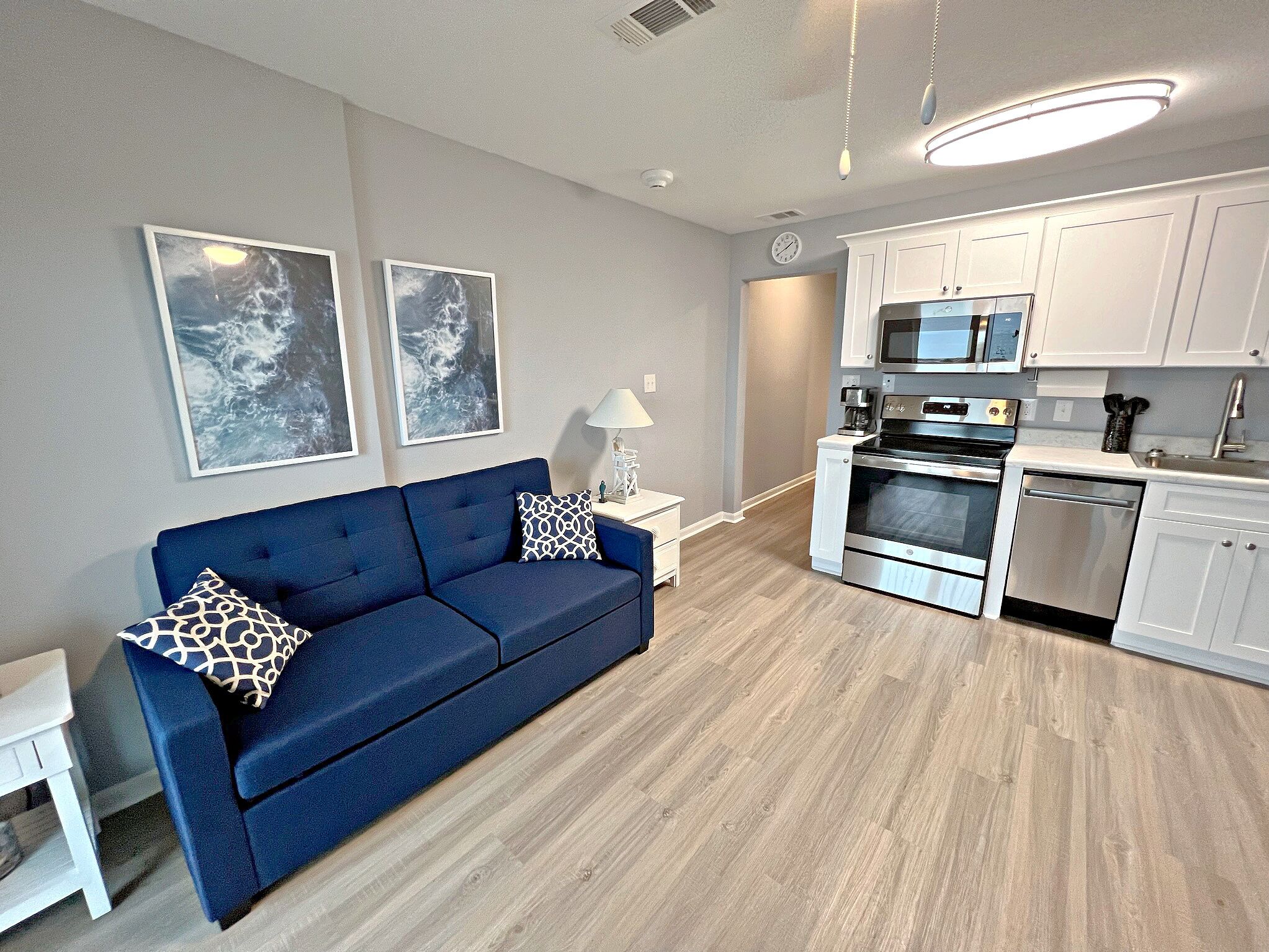 A modern living room with a blue sofa, a white side table, and a lamp. Adjacent is a kitchen with white cabinets, a microwave, an oven, and a dishwasher.