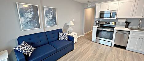 A modern living room with a blue sofa, a white side table, and a lamp. Adjacent is a kitchen with white cabinets, a microwave, an oven, and a dishwasher.