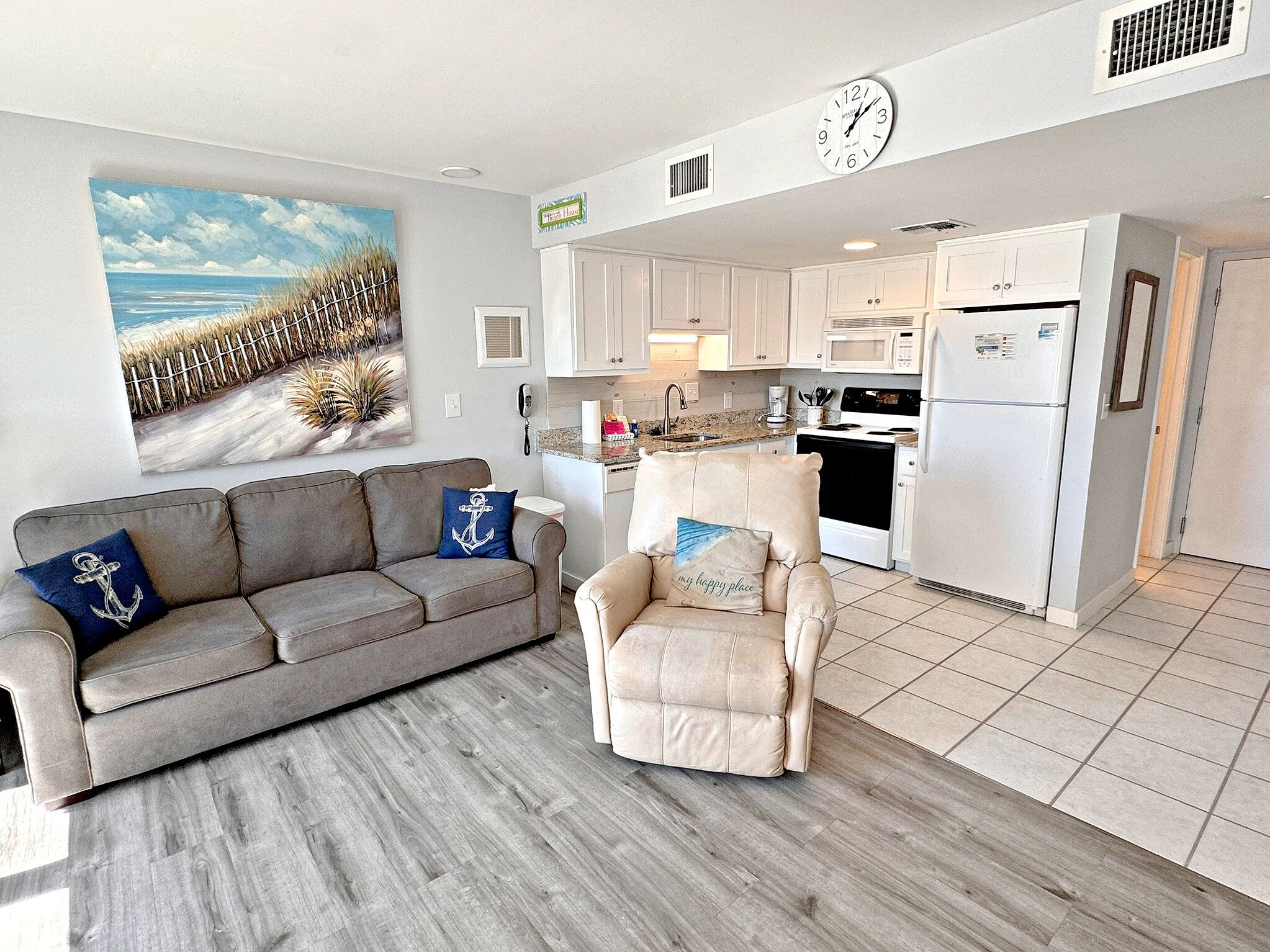 A living room with a grey sofa and armchair, a painting of a beach, and a kitchen with white cabinets and appliances.