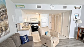 A living room with a beige sofa and armchair, a kitchen in the background, and a hallway leading to a door. - Gulf Shores Plantation IV (Gulf Shores)