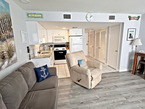 A living room with a beige sofa and armchair, a kitchen in the background, and a hallway leading to a door. - Gulf Shores Plantation IV (Gulf Shores)