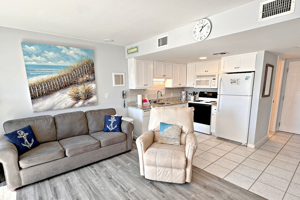 A living room with a grey sofa and armchair, a painting of a beach, and a kitchen with white cabinets and appliances.