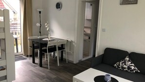 Een moderne woonkamer met een zwarte bank, een witte salontafel en een eettafel met witte stoelen. Er hangt een klok aan de muur en een bord met Duitse tekst.