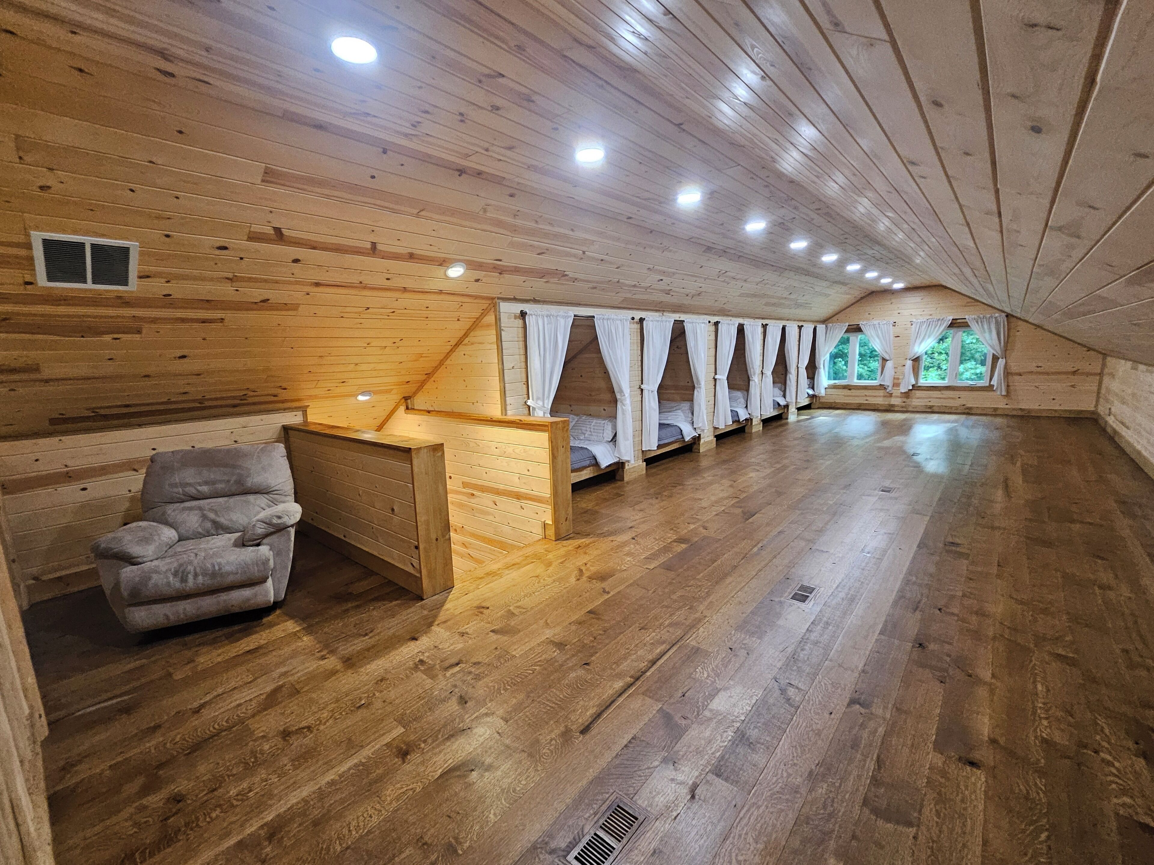 A wooden interior with a row of beds, a cushioned chair, and a small window.