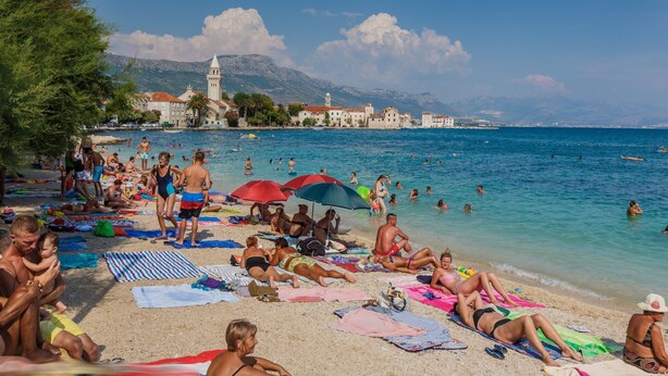 Praia lotada com pessoas tomando sol e nadando, céu azul límpido e um edifício histórico ao fundo.