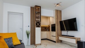 Uma sala de estar moderna com um sofá, uma TV de tela plana e uma cozinha visível por uma porta aberta.