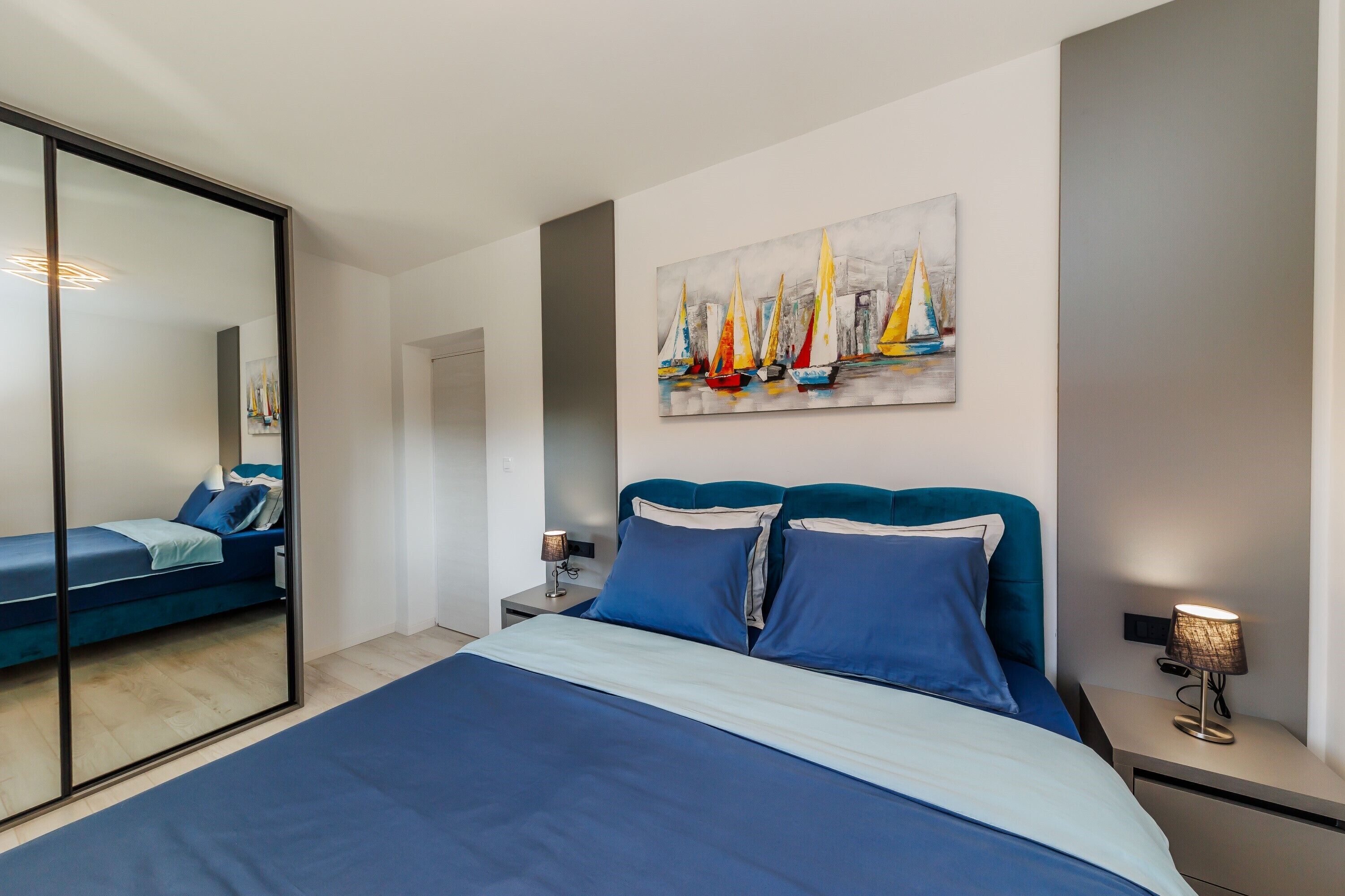 A modern bedroom with a bed, a nightstand, a lamp, and a painting of sailboats on the wall.