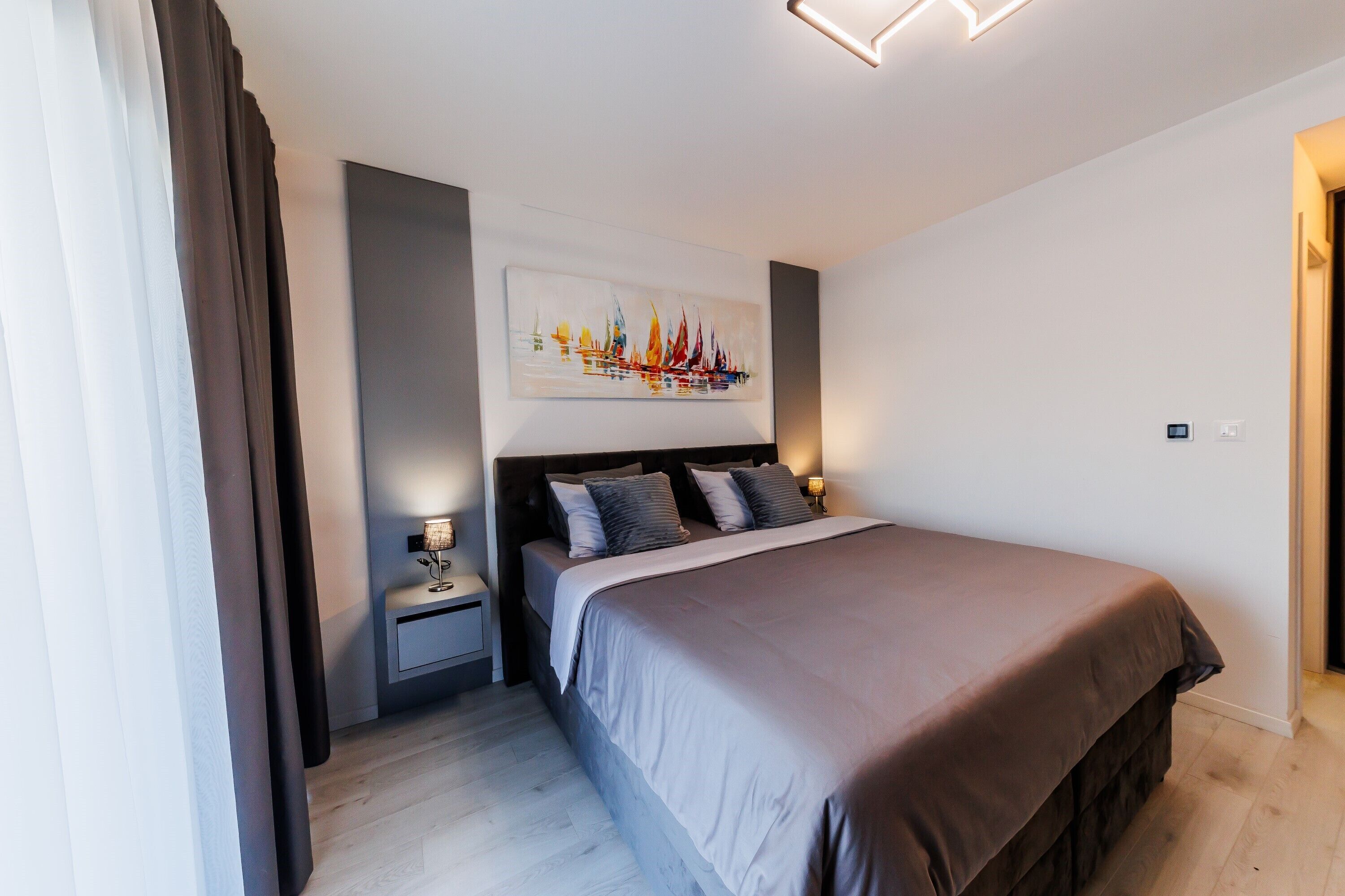 A modern bedroom with a large bed, a nightstand, a wall-mounted painting, and a ceiling light fixture.