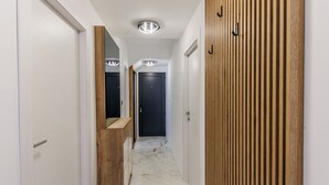 Corredor com paredes brancas, painel de madeira em uma lateral e chão de mármore. Há portas e um banco de madeira.