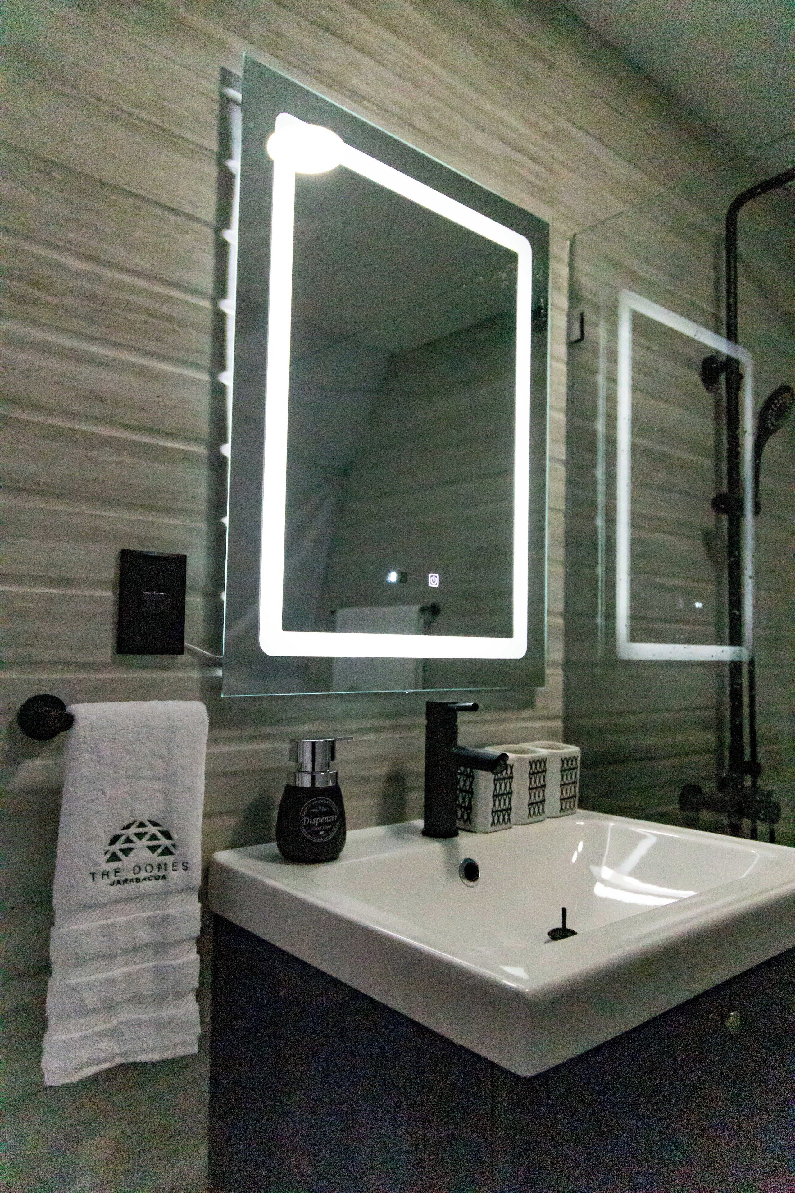 A bathroom with a white sink, a mirror with a light, a towel, and a soap dispenser.