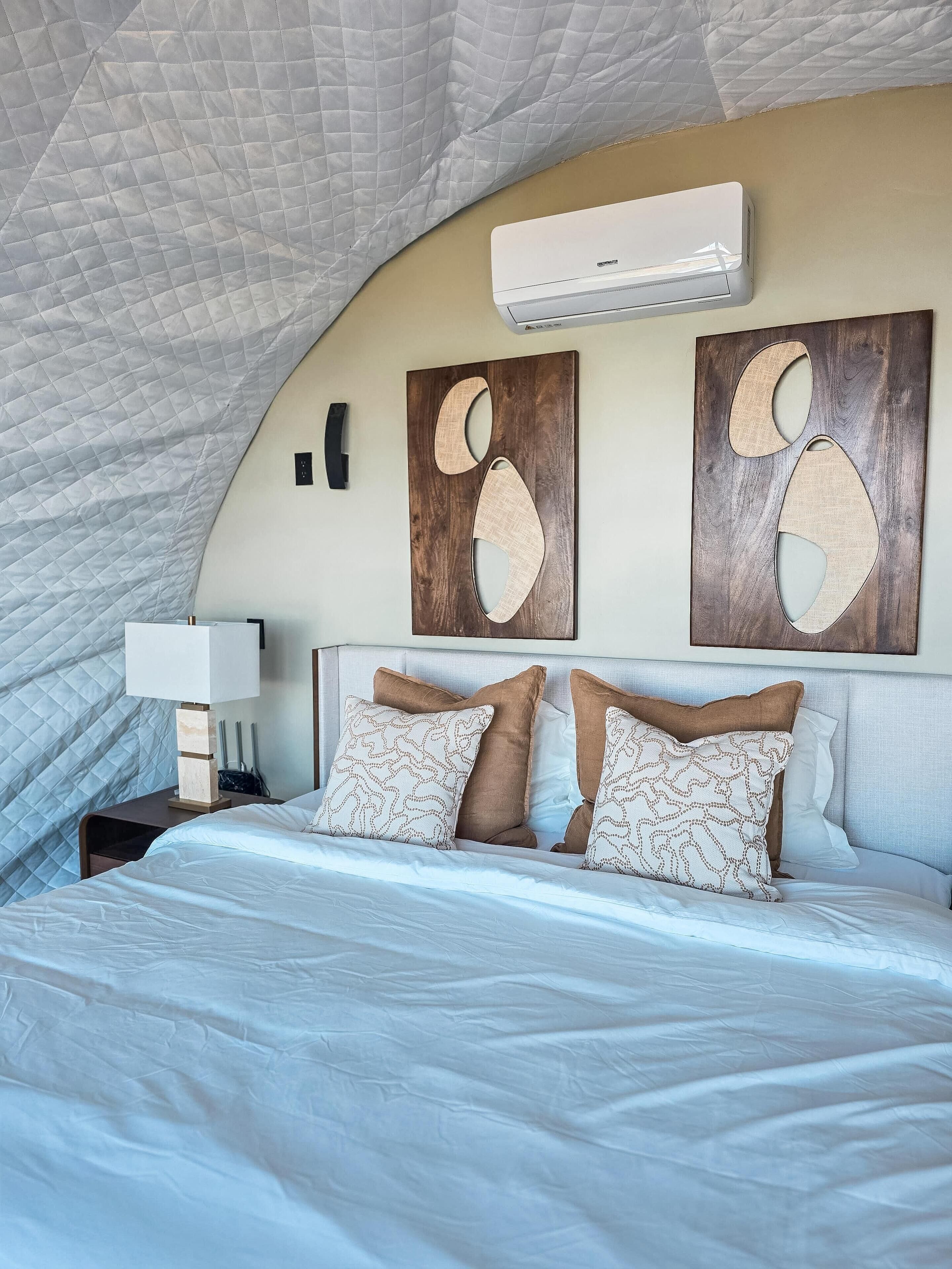 Une chambre à coucher avec un grand lit, une table de chevet, un climatiseur mural et des œuvres d’art abstraites accrochées au mur.