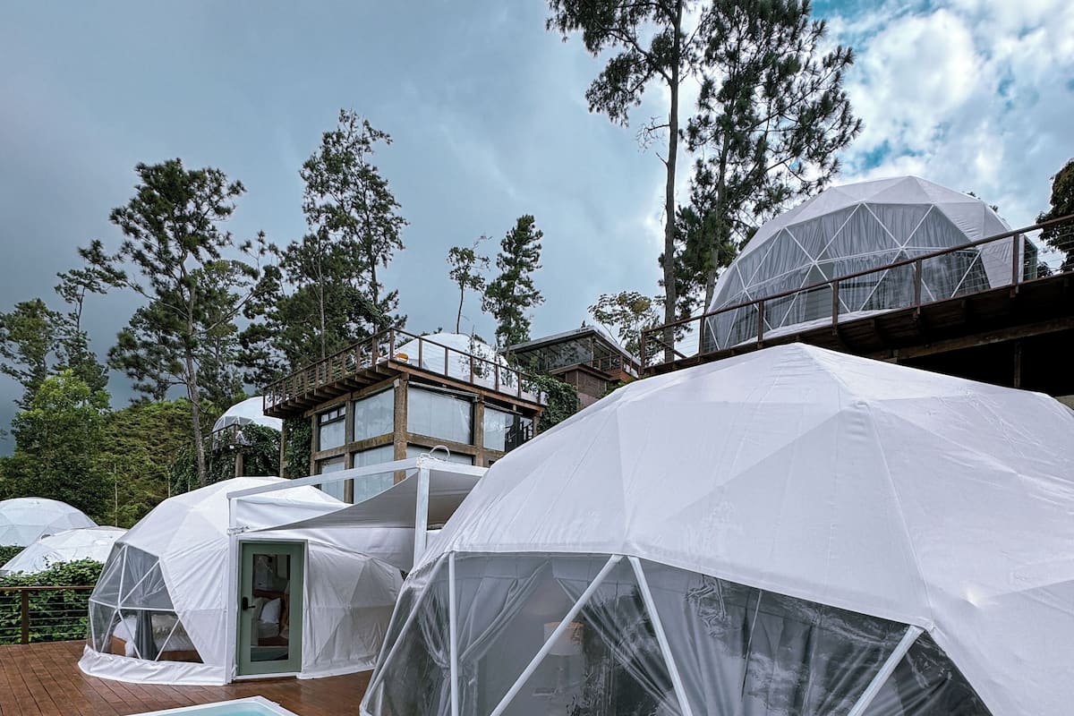 Hébergements en forme de dôme géodésique avec une piscine sur une terrasse en bois, entourés d’arbres et sous un ciel nuageux.