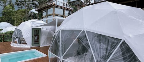 Alojamiento en cúpula geodésica con piscina en terraza de madera, rodeado de árboles y un cielo nublado.