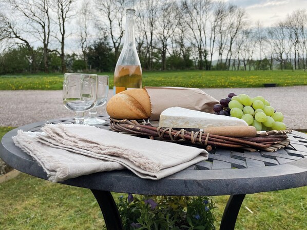 Una mesa al aire libre con una manta, una botella, pan, queso, uvas y otros alimentos.