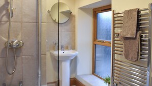 A bathroom with a glass shower enclosure, a pedestal sink, a round mirror, a towel rack, and a window. - Pippa's Cottage - UK42332 (Greystoke, near Penrith)