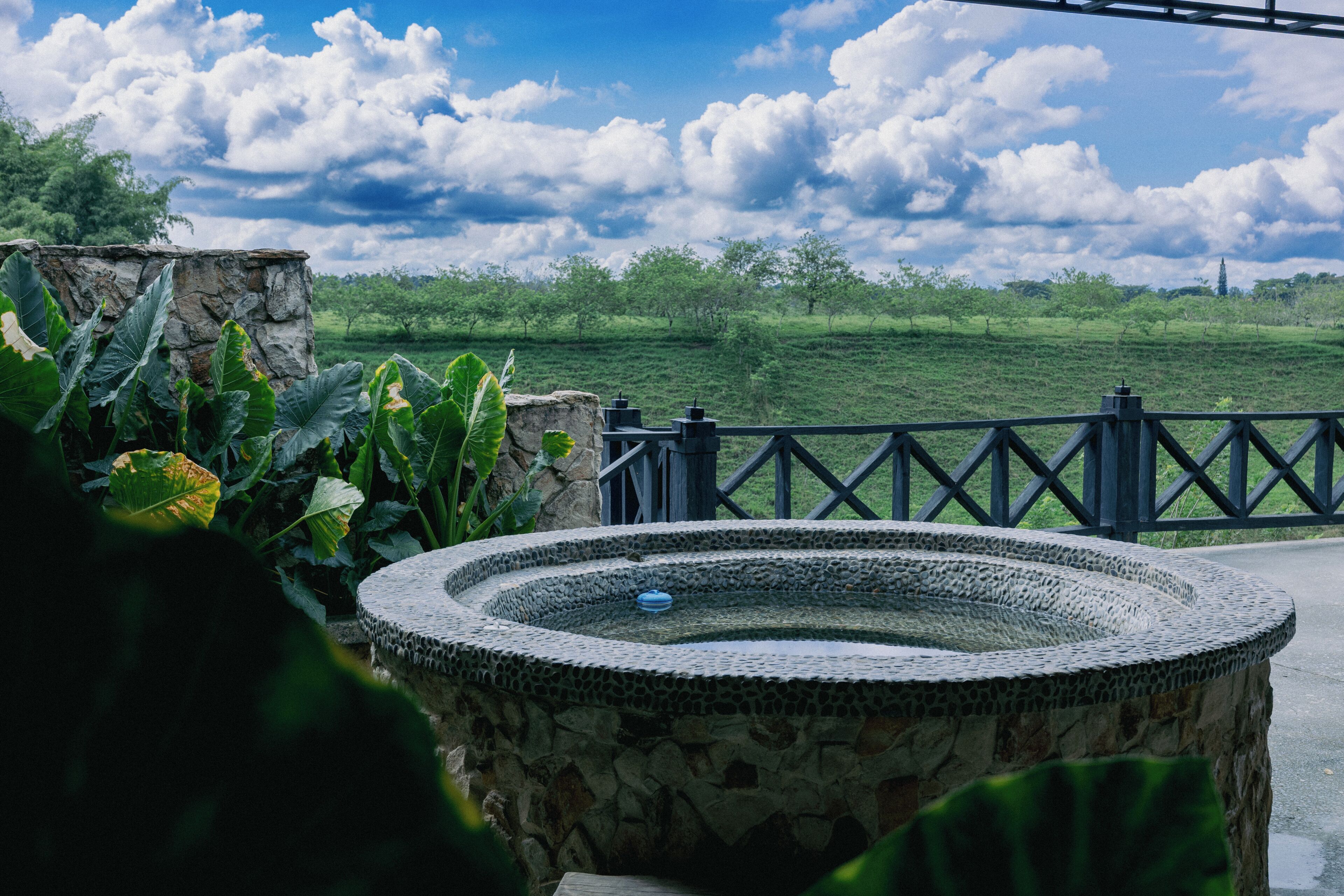 Une fontaine en pierre agrémentée d’une rambarde métallique, entourée de verdure et offrant une vue sur un champ herbeux sous un ciel partiellement nuageux.