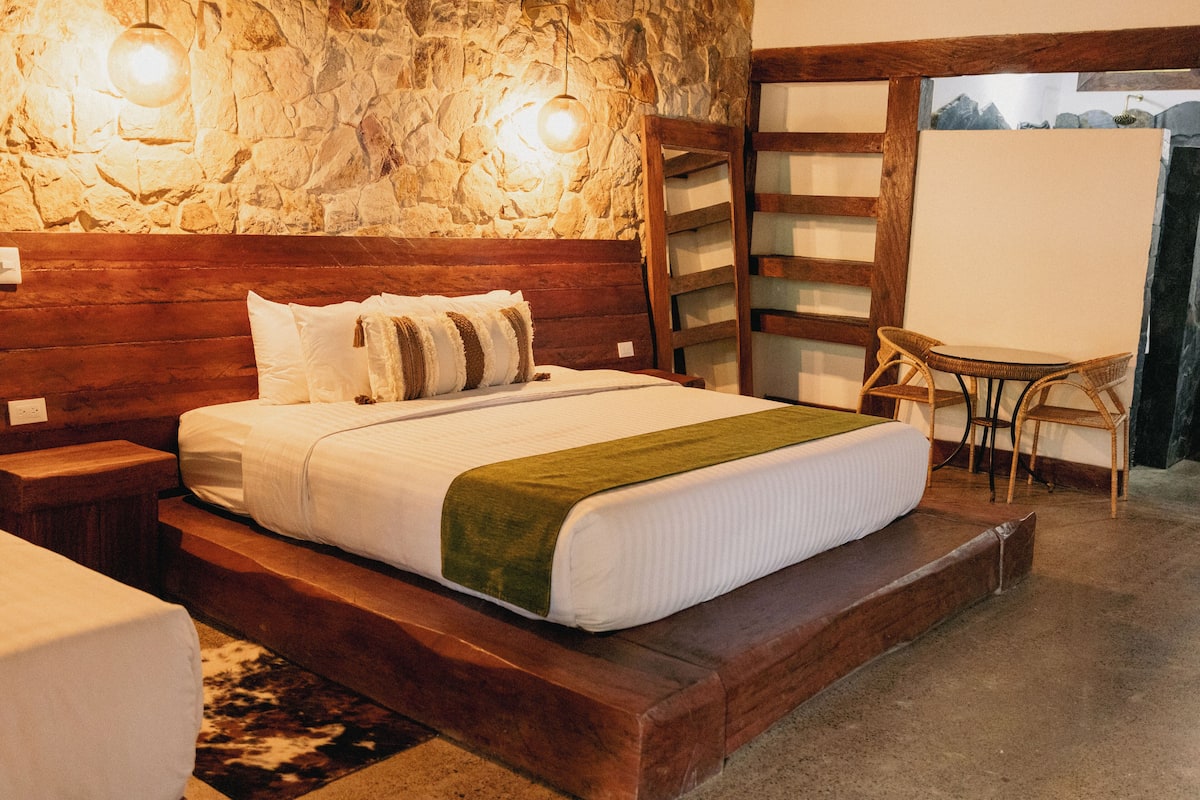 Une chambre avec un mur en pierre, une tête de lit en bois, un lit avec une literie blanche, un plaid vert, une table de chevet, une chaise et une petite table.