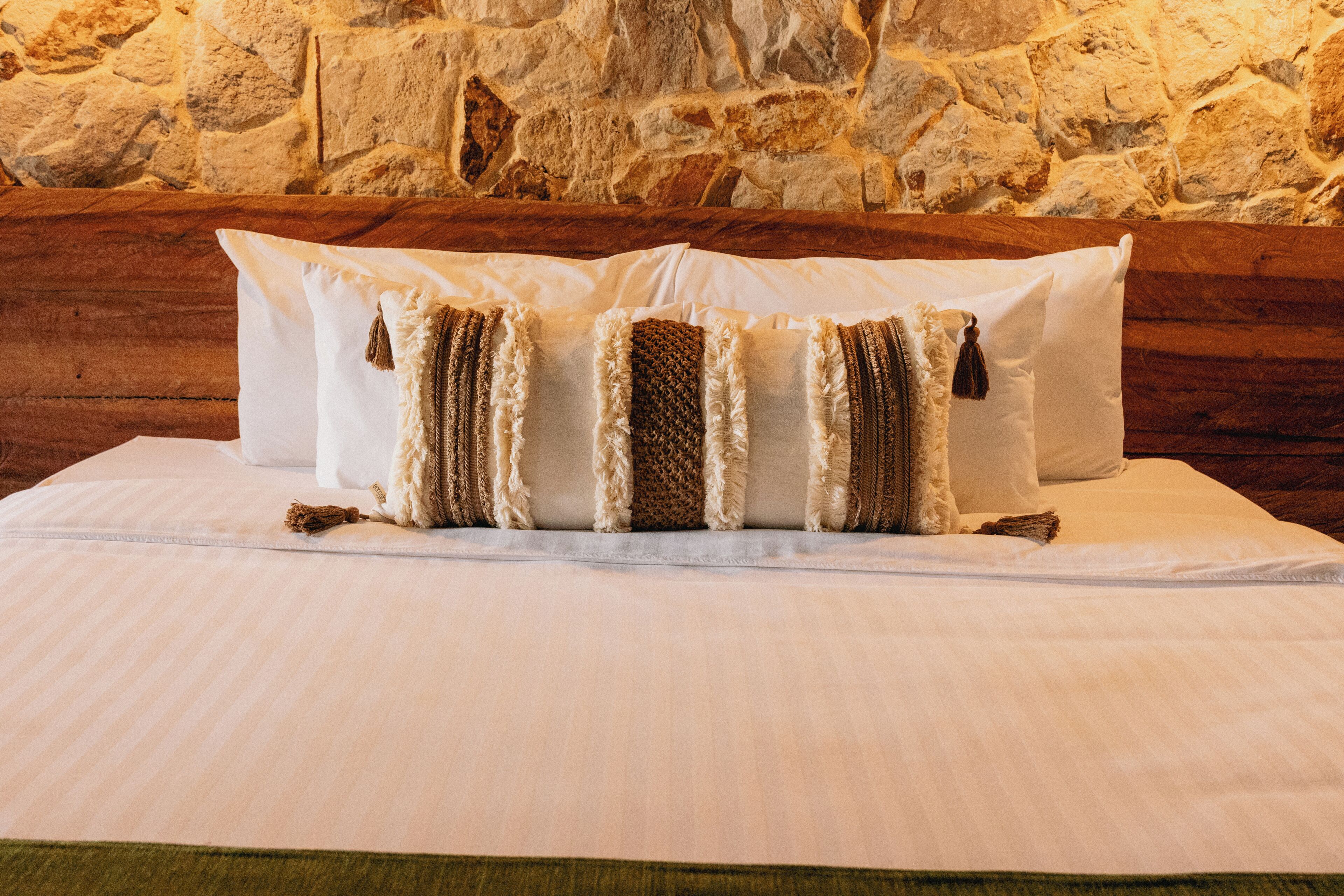 A bed with white linens and striped pillows against a stone wall.
