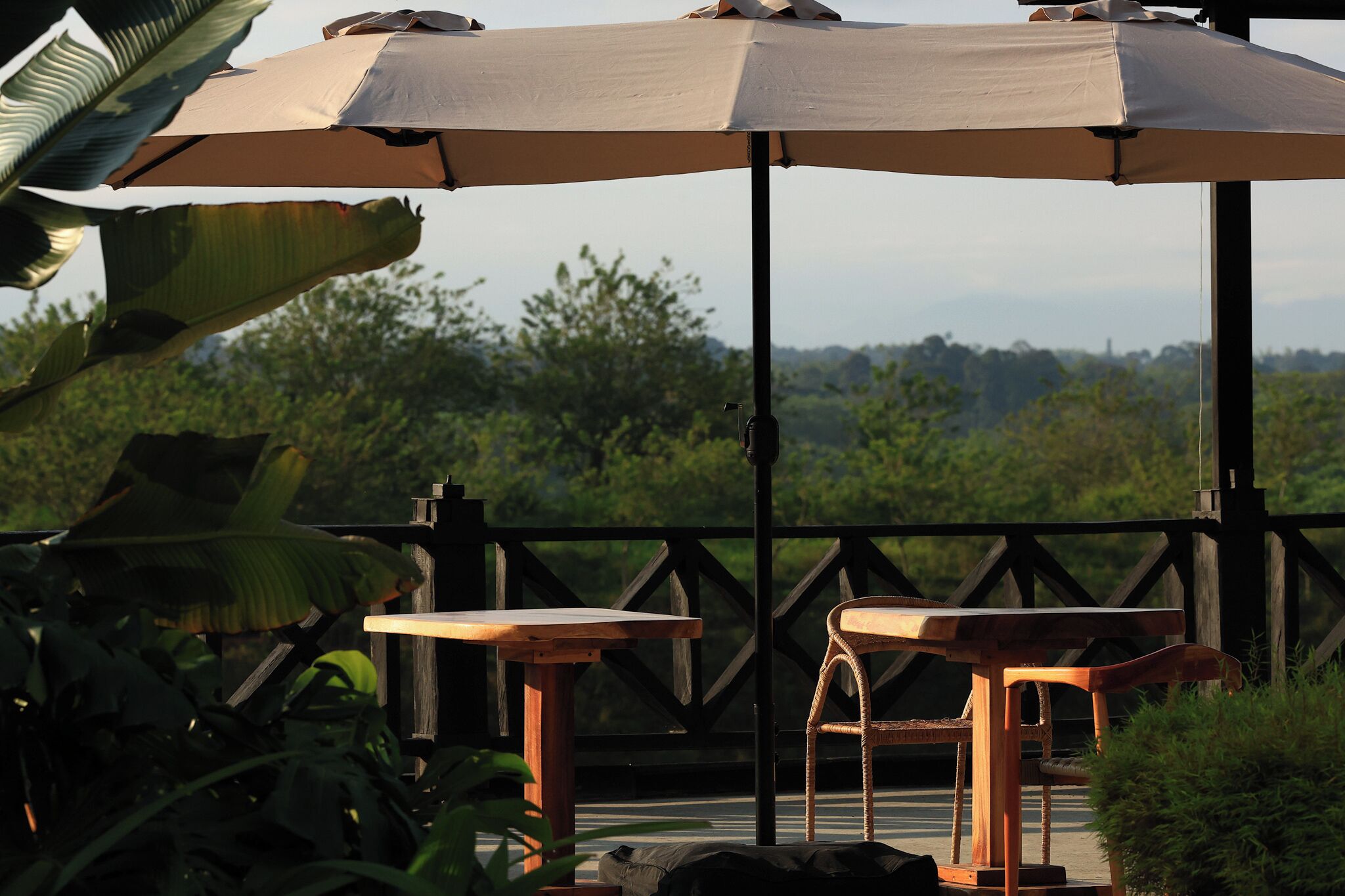 A wooden deck with two tables and an umbrella, surrounded by lush greenery and a scenic view.
