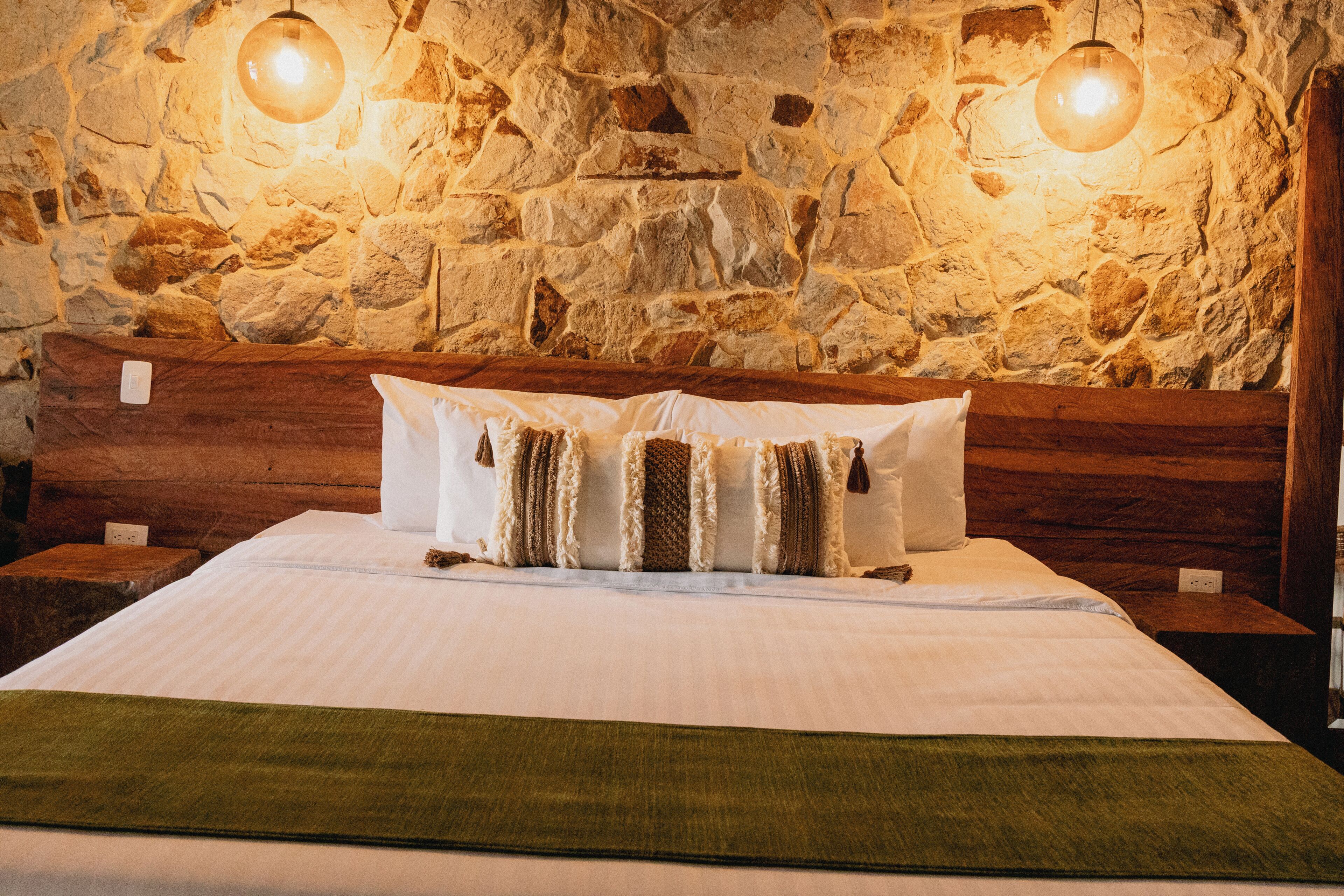 A bed with a wooden headboard, a stone wall, and two wall-mounted lamps.