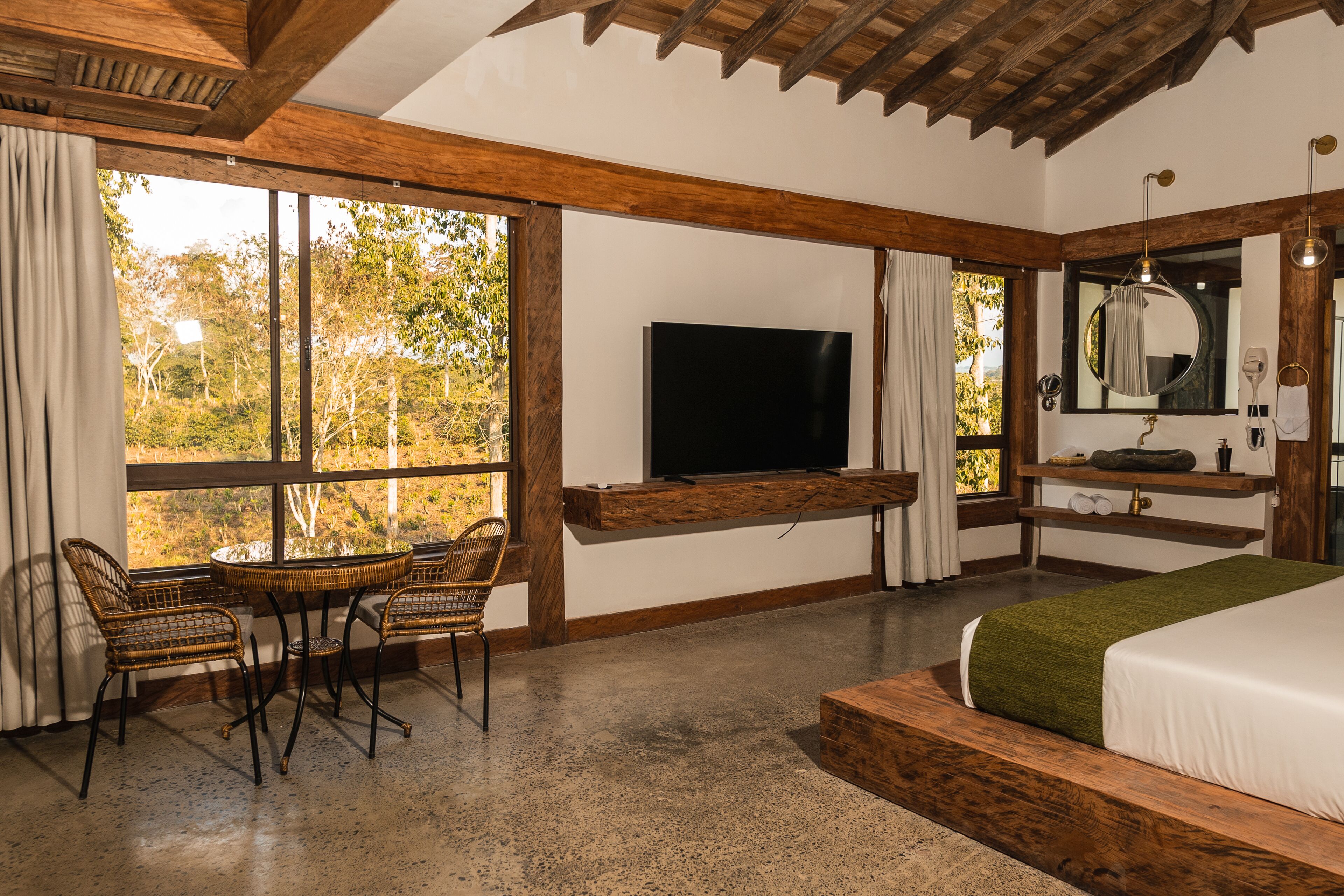 A bedroom with a bed, a television, a seating area with chairs, and a window with a view of trees.