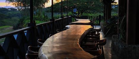 Una larga mesa de madera con sillas bajo una zona techada, con vista a un paisaje exuberante.