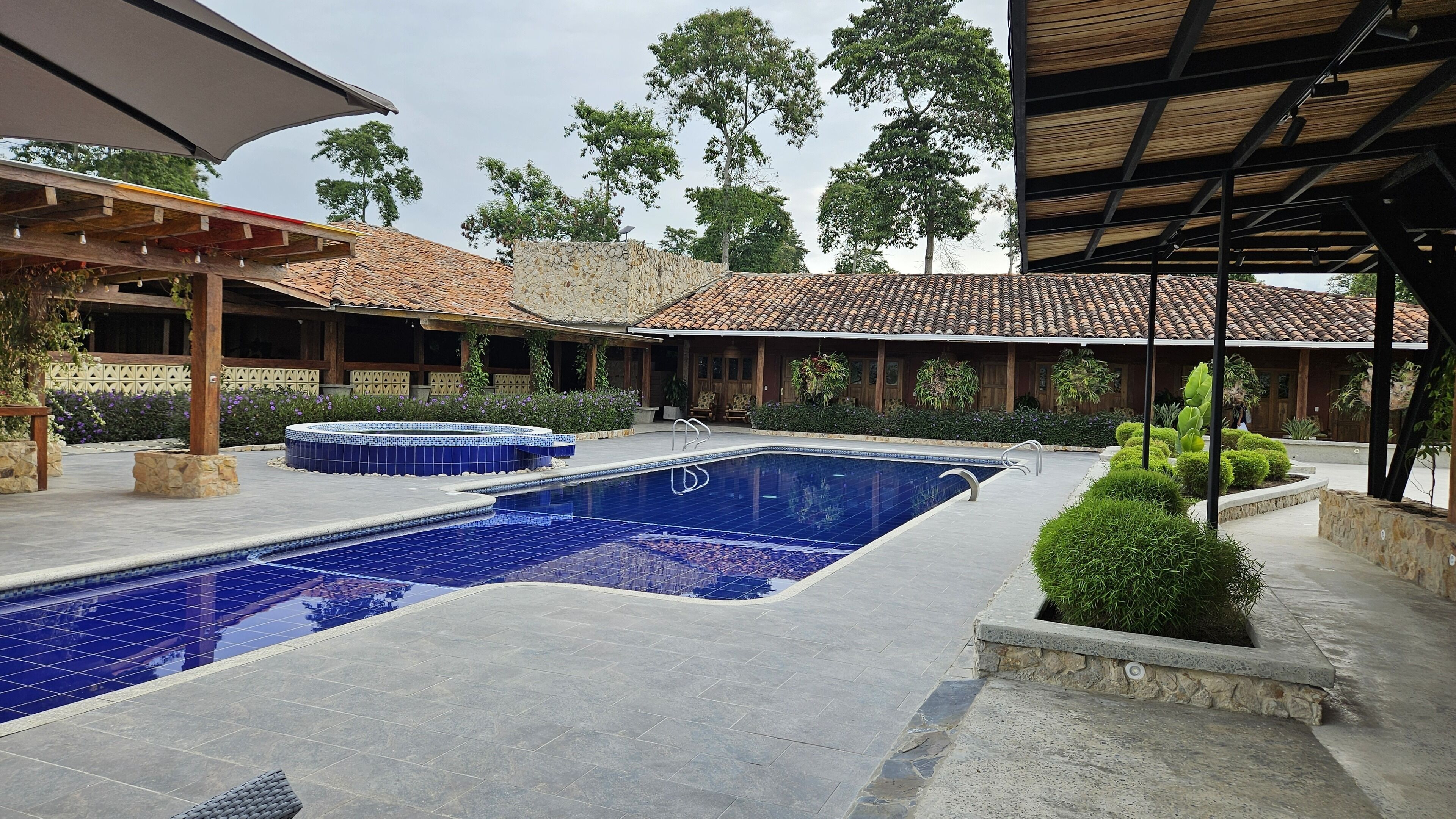 A swimming pool with a tiled deck, surrounded by stone walls and covered areas with wooden beams.