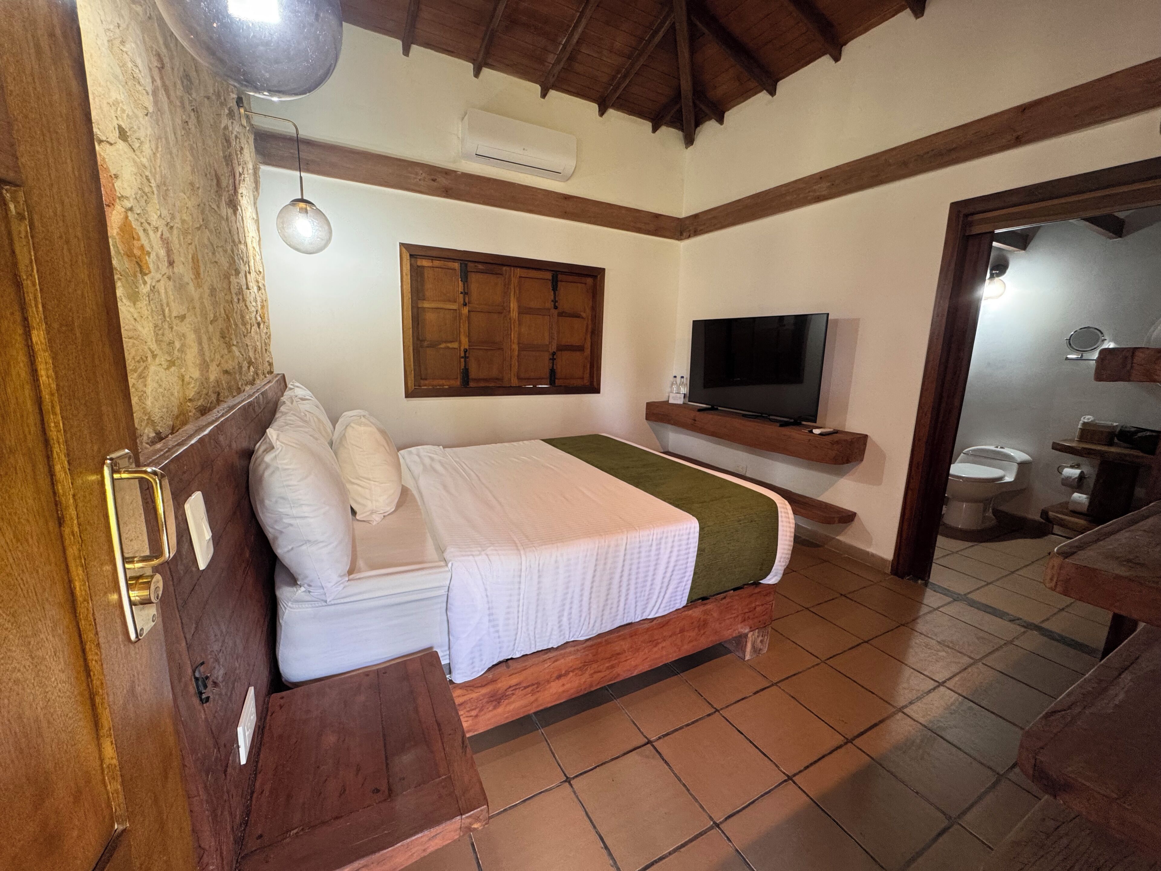 Un dormitorio con cama, televisión, techo de madera y un baño visible a través de una puerta abierta.