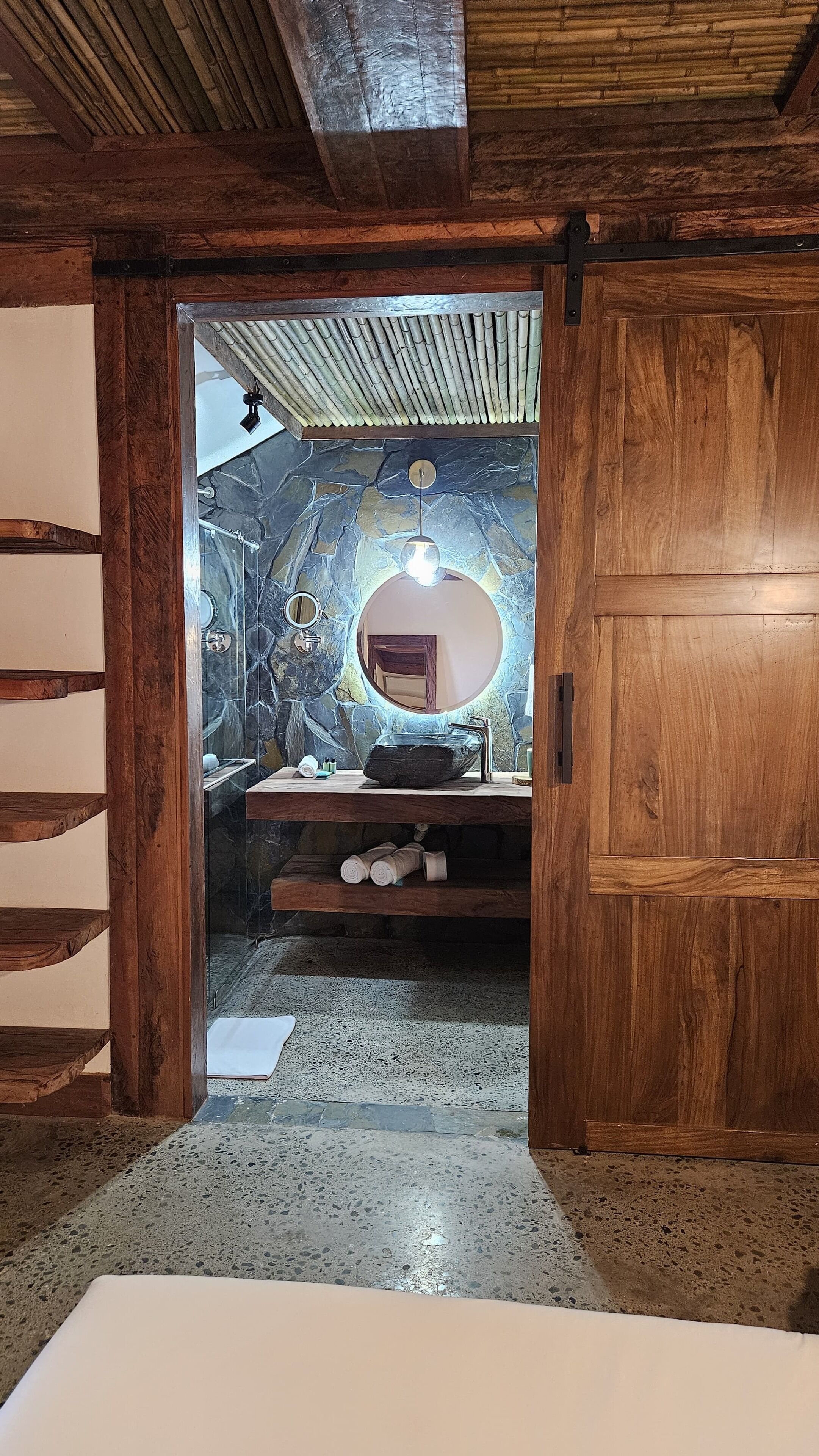 A bathroom with a stone wall, a round mirror, and a wooden door.