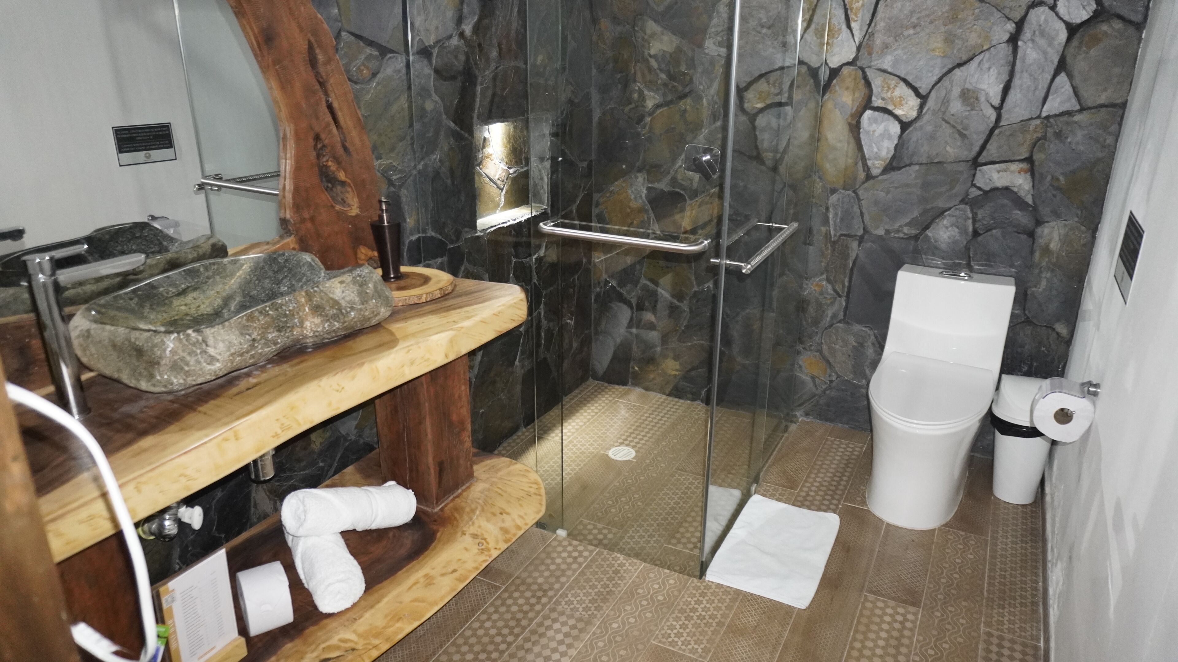 A bathroom with a stone wall, a wooden shelf with a basin, a glass shower enclosure, and a toilet.