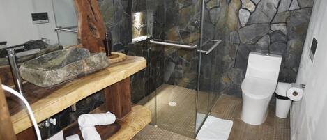A bathroom with a stone wall, a wooden shelf with a basin, a glass shower enclosure, and a toilet.