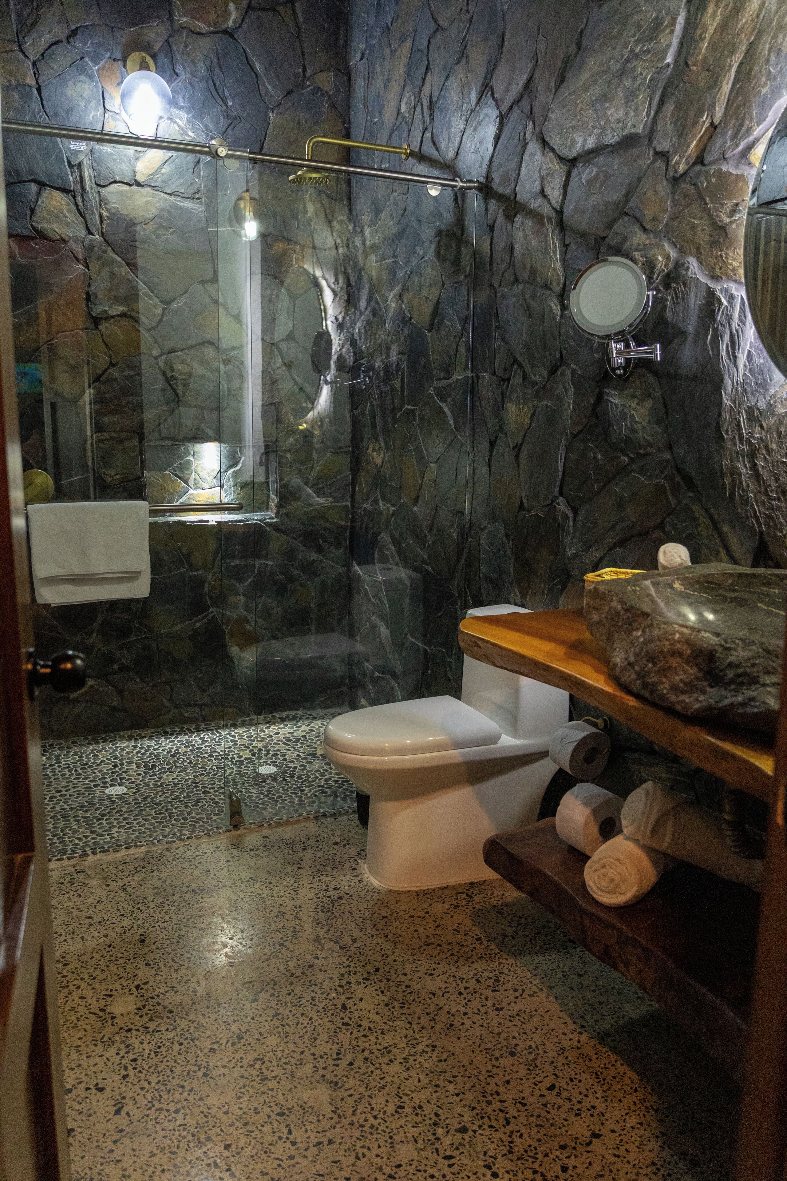 A bathroom with a stone wall, a glass shower enclosure, a toilet, and a wooden vanity with a sink.